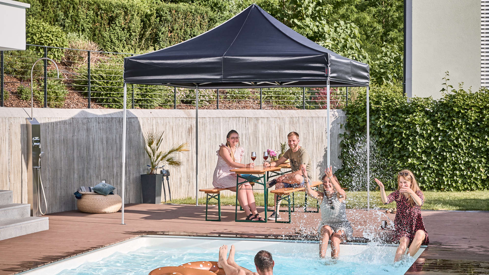 In the garden, the black 3x3 m folding canopy tent and classic beer garden table set. Friends spend the day in the garden and the pool.