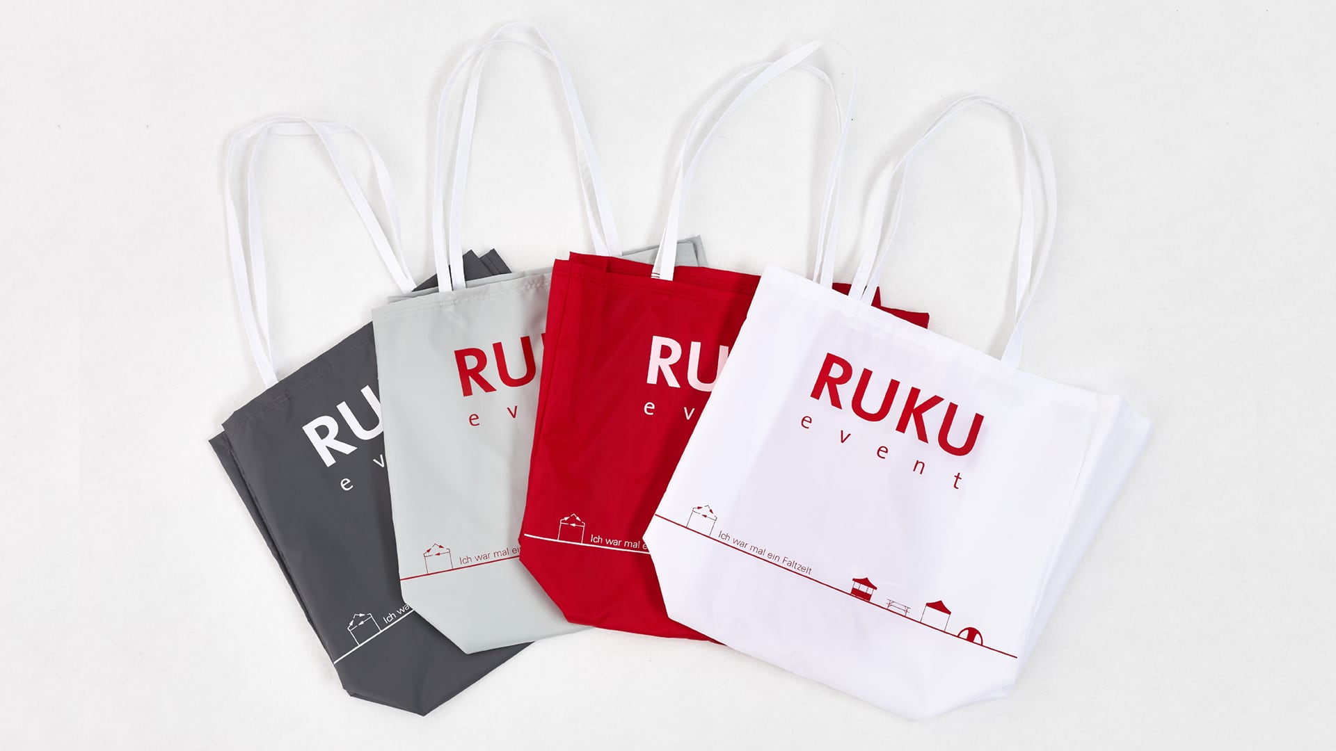 Die nachhaltigen RUKUevent Einkaufstaschen liegen nebeneinander überlappend in den Farben dunkelgrau, hellgrau, rot und weiß mit aufgenähtem RUKUevent Logo.