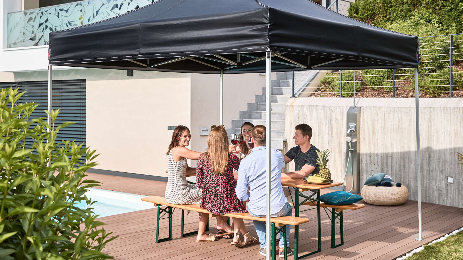 In the garden, the black 3x3 m folding canopy tent and classic beer garden table set. Friends spend the day in the garden in front of the pool.