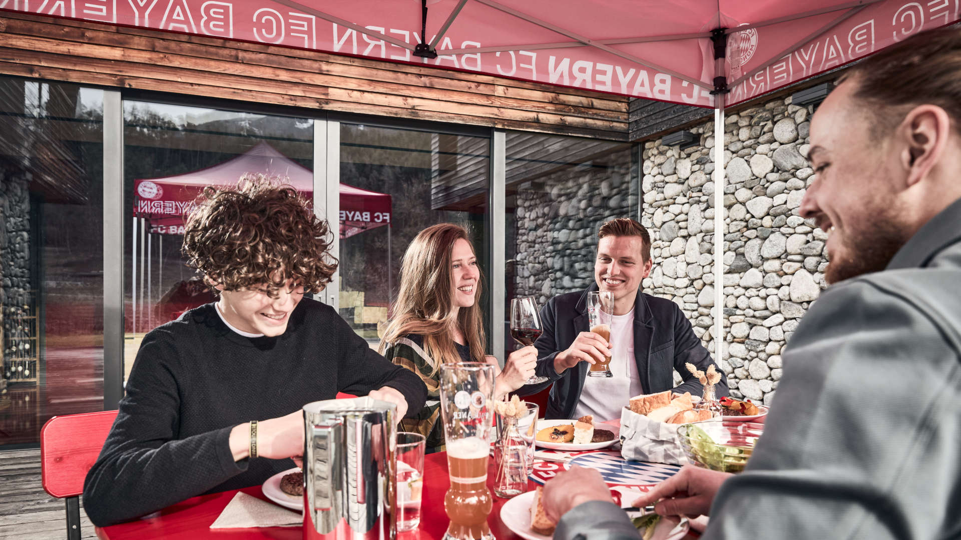 Freunde genießen auf der Bierzeltgarnitur mit Lehne unter dem FC Bayern München Faltpavillon gemeinsam das Essen.