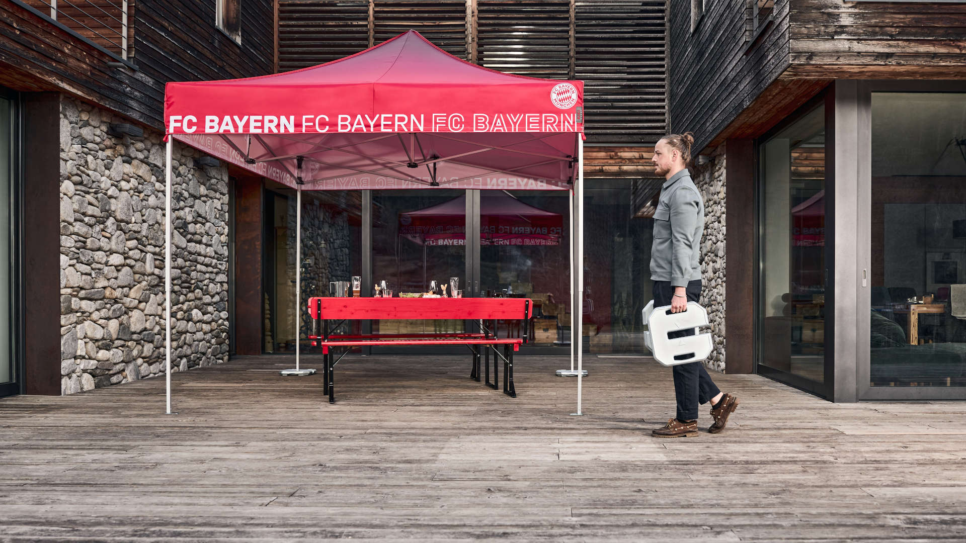 Ein Mann bringt die 25 kg schweren Bodenplatten zum roten FC Bayern München Faltpavillon. Unter dem Zelt ist die rote FCB Bierzeltgarnitur bereits gedeckt.