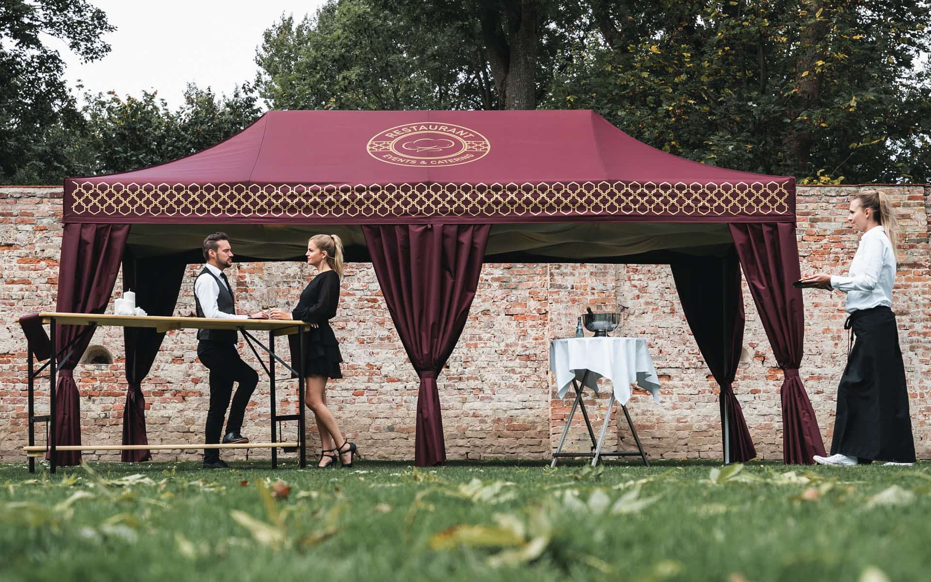 Zwei elegant gekleidete Gäste warten an einem Stehtisch vor dem Bordeaux-Roten Faltpavillon mit Eckvorhängen auf die Kellnerin, die ihre Drinks serviert.